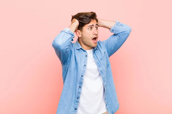 young handsome man with open mouth, looking horrified and shocked because of a terrible mistake, raising hands to head against pink background