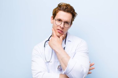 Genç kızıl saçlı doktor ciddi, düşünceli ve güvensiz görünüyor, bir kolu çapraz ve elleri çenesinde, mavi duvara karşı seçenekleri tartıyor.
