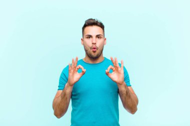 Genç İspanyol adam şok olmuş, şaşırmış ve şaşırmış hissediyor. Onaylıyor. İki eliyle mavi arka plana işaret koyuyor.