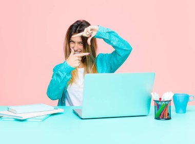 Bilgisayarla çalışan genç ve güzel bir kadın mutlu, arkadaş canlısı ve pozitif hissediyor, gülümsüyor ve elleriyle bir portre veya fotoğraf çerçevesi yapıyor.