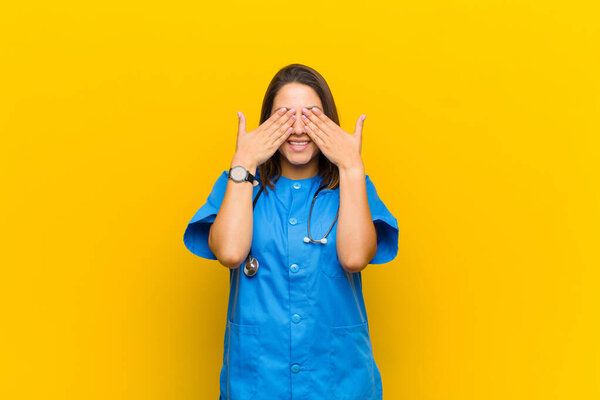 smiling and feeling happy, covering eyes with both hands and waiting for unbelievable surprise isolated against yellow wall