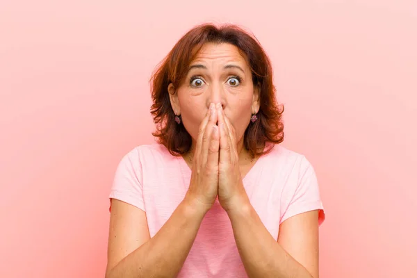 middle age woman feeling worried, upset and scared, covering mouth with hands, looking anxious and having messed up against pink wall