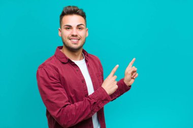 Genç İspanyol adam mutlu bir şekilde gülümsüyor ve her iki elinde fotokopi uzayında mavi arka planda nesne gösteriyor.