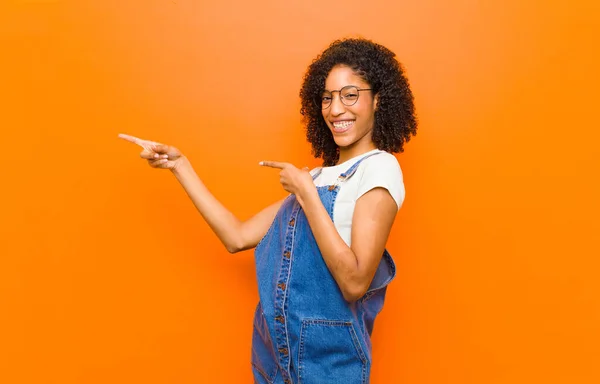 年轻漂亮的黑人女人感到快乐和惊奇 带着震惊的表情微笑着 指指旁边的橙色墙壁 — 图库照片