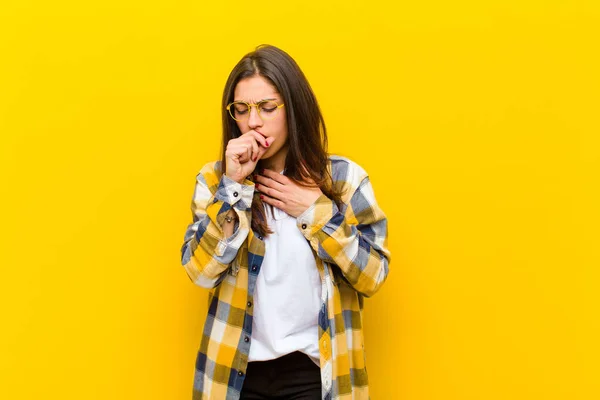 young  pretty woman feeling ill with a sore throat and flu symptoms, coughing with mouth covered against orange wall