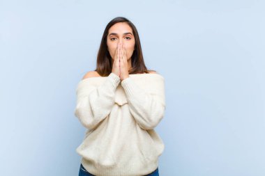 Genç, güzel bir kadın mutlu ve heyecanlı, şaşırmış ve hayrete düşmüş elleriyle ağzını kapatmış, mavi arka planda sevimli bir ifadeyle kıkırıyor.