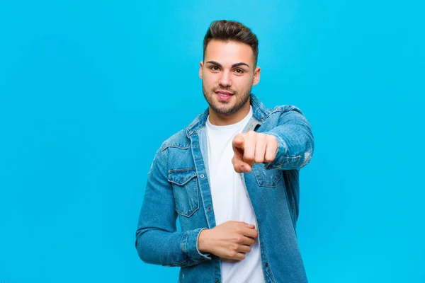 young hispanic man pointing at camera with a satisfied, confident, friendly smile, choosing you against blue background