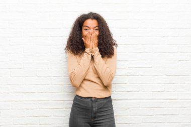 Genç siyahi kadın mutlu, neşeli, şanslı ve şaşkın görünüyor. İki eli tuğla duvara dayalı ağzını kapatıyor.