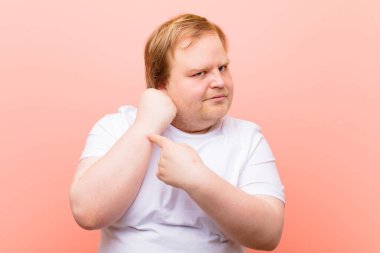 Sabırsız ve sinirli görünen, saati gösteren, dakiklik isteyen genç, iri yarı bir adam, pembe duvara karşı zamanında olmak istiyor.