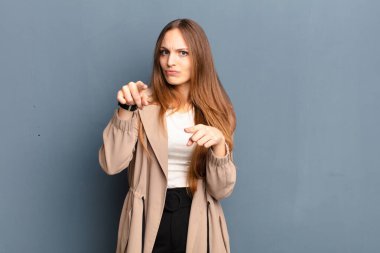 Genç ve güzel bir kadın kameraya hem parmaklarını hem de kızgın yüzünü göstererek gri arka plana karşı görevini yapmanı söylüyor.