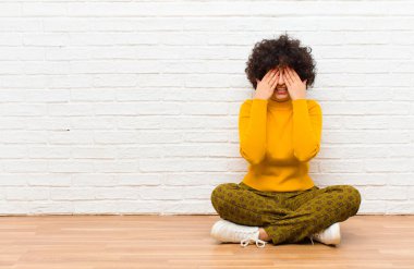 Genç, güzel afro kadın gülümseyen ve mutlu hisseden, iki eliyle gözlerini kapayan ve yerde oturan inanılmaz sürprizi bekleyen