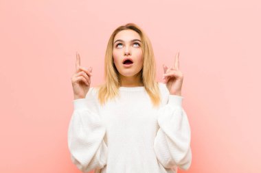 Şok olmuş, şaşkın ve ağzı açık, pembe düz duvara uzayı kopyalamak için iki elini havaya kaldırmış genç, güzel sarışın kadın.