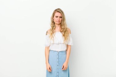 Genç, sarışın bir kadın üzgün ve mızmız görünüyor. Mutsuz bir bakışla ağlıyor. Beyaz duvara karşı olumsuz ve hayal kırıklığına uğramış bir tavırla ağlıyor.