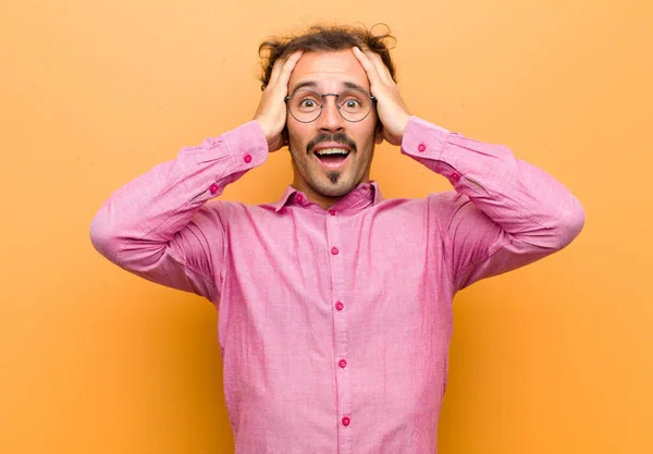 young handsome man raising hands to head, open-mouthed, feeling extremely lucky, surprised, excited and happy against orange wall