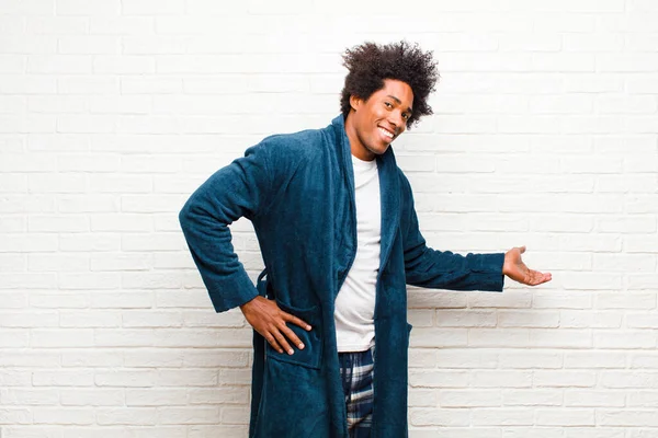 young black man wearing pajamas with gown feeling happy and cheerful, smiling and welcoming you, inviting you in with a friendly gesture against brick wall