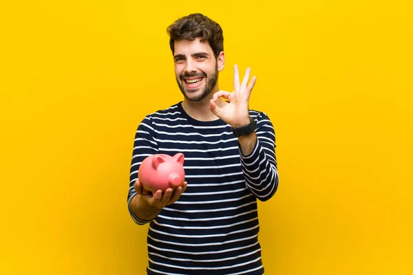 Ung stilig man som håller en spargris mot orange backgroun — Stockfoto