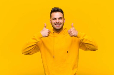 Genç İspanyol adam gülümsüyor. Mutlu, pozitif, kendinden emin ve başarılı görünüyor. İki başparmağı da turuncu duvarda.