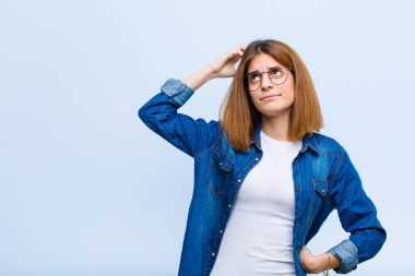 Genç, güzel bir kadın şaşkın ve kafası karışmış hissediyor. Kafasını kaşıyor ve mavi arka plana bakıyor.