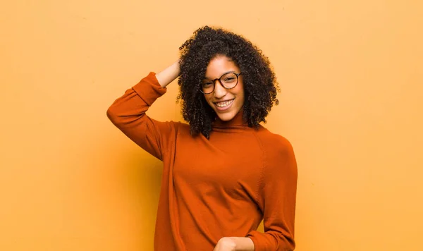 young pretty black woman smiling cheerfully and casually, taking hand to head with a positive, happy and confident look against orange wall