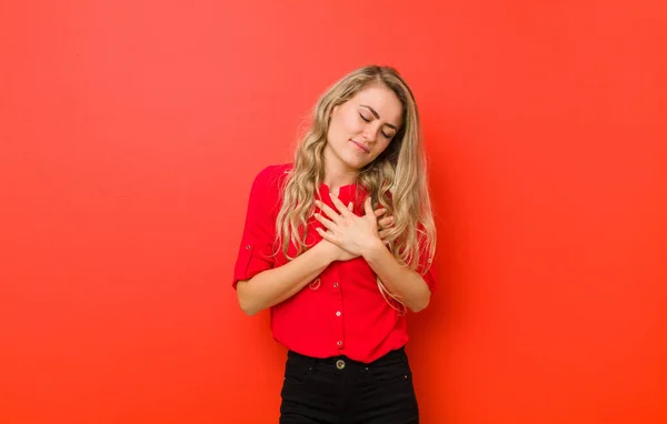 若いブロンドの女性は悲しく 傷つき 胸を痛め 両手を心の近くに持ち 赤い壁に対して落ち込んでいる — ストック写真
