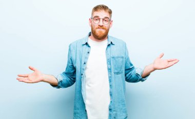 Genç kızıl saçlı adam şaşkın ve şaşkın hissediyor, doğru cevap ya da karardan emin değil, yumuşak mavi arka plana karşı bir seçim yapmaya çalışıyor.