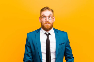 Genç kızıl saçlı işadamı aptal ve komik görünüyor, şaşı bir ifadeyle, şakalaşıyor ve turuncu arka planda takılıyor.