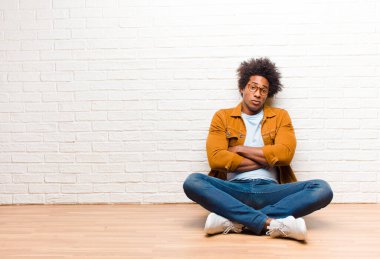 Genç siyahi bir adam hayal kırıklığına uğramış, hayal kırıklığına uğramış, ciddi görünüyor, kızgın ve kızgın kollarını kavuşturup evde yerde oturuyor.