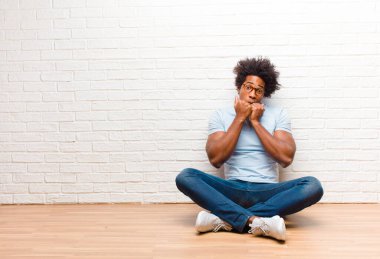 Genç siyah adam endişeli, gergin, stresli ve korkmuş görünüyor, tırnaklarını yiyor ve evde yerde oturan yanal fotokopi odasına bakıyor.