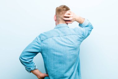 Genç kızıl saçlı adam düşünüyor ya da şüphe ediyor, kaşınıyor, kafası karışık ve şaşkın hissediyor, arka ya da arka plan yumuşak mavi arka plana karşı