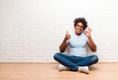 Genç siyahi adam mutlu, şaşkın, tatmin olmuş ve şaşırmış hissediyor, tamam gösteriyor ve parmaklarını kaldırıyor, gülümsüyor evde yerde oturuyor.