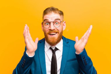 Genç kızıl saçlı işadamı turuncu arka planda beklenmedik bir sürprizden dolayı şaşkın ve heyecanlı, güler yüzlü, şaşkın ve mutlu hissediyor.