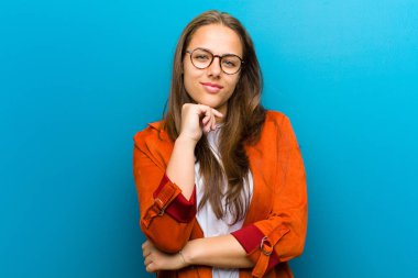 Genç bir kadın ciddi, düşünceli ve güvensiz görünüyor, bir kolu çapraz ve çene üzerinde, mavi arka planda seçenekleri tartıyor
