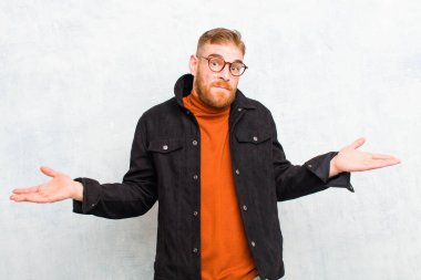 Genç kızıl saçlı adam şaşkın ve şaşkın hissediyor, doğru cevap veya karardan emin değil, grunge duvarına karşı bir seçim yapmaya çalışıyor.