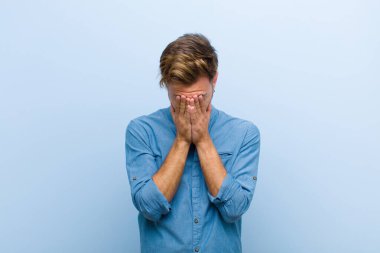 Genç işadamı üzgün, hayal kırıklığına uğramış, gergin ve depresif hissediyor, iki eliyle yüzünü kapatıyor, mavi arka planda ağlıyor.