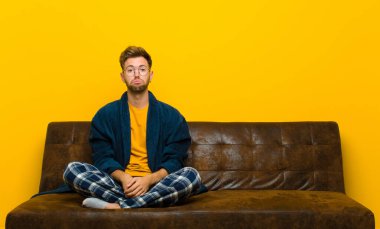 Genç adam pijama giyiyor üzgün ve mızmızlanıyor mutsuz bir bakışla, negatif ve hayal kırıklığına uğramış bir tavırla ağlıyor. Kanepede oturuyor.