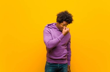 Genç siyah adam stresli, mutsuz ve hayal kırıklığına uğramış, alnına dokunuyor ve turuncu duvara karşı şiddetli baş ağrısı çekiyor.