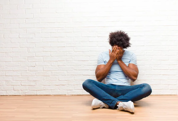 young black man feeling sad, frustrated, nervous and depressed, covering face with both hands, crying sitting on the floor at home
