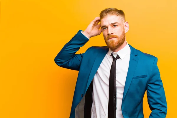 young red head businessman feeling puzzled and confused, scratching head and looking to the side against orange background