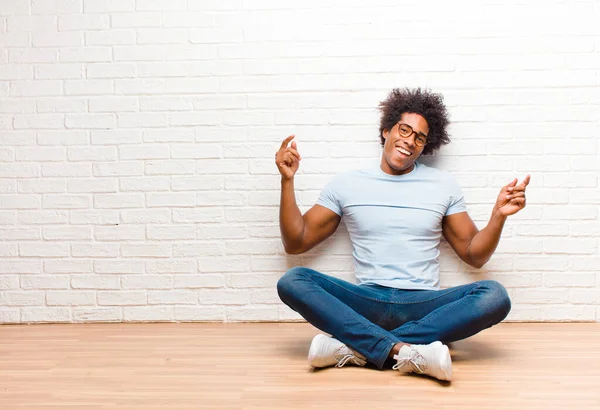 young black man smiling, feeling carefree, relaxed and happy, dancing and listening to music, having fun at a party sitting on the floor at home