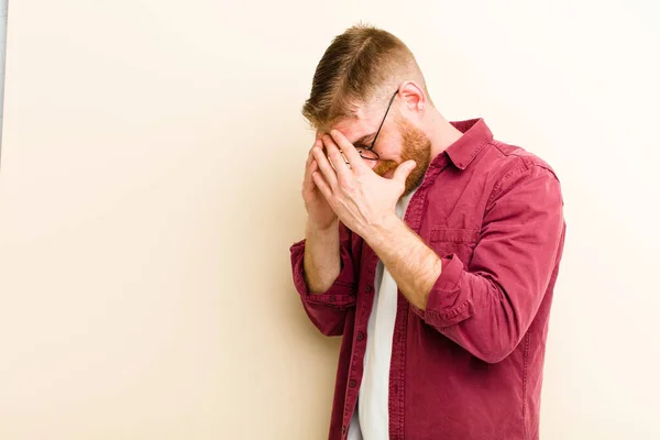 young red head man covering eyes with hands with a sad, frustrated look of despair, crying, side view against beige background