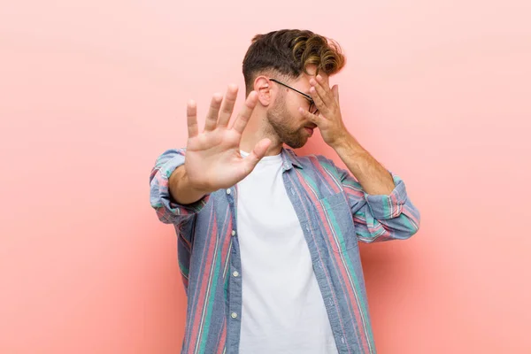 young man covering face with hand and putting other hand up front to stop camera, refusing photos or pictures against pink background