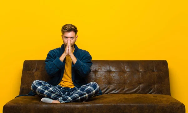 young man wearing pajamas feeling worried, hopeful and religious, praying faithfully with palms pressed, begging forgiveness . sitting on a sofa