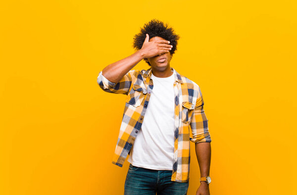 Young black man covering eyes with one hand feeling scared or anxious, wondering or blindly waiting for a surprise against orange wall