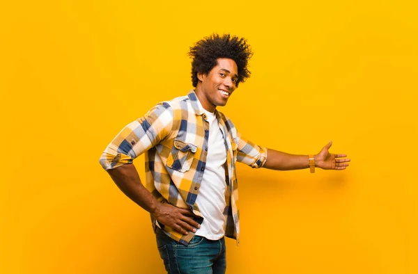 young black man feeling happy and cheerful, smiling and welcoming you, inviting you in with a friendly gesture against orange wall