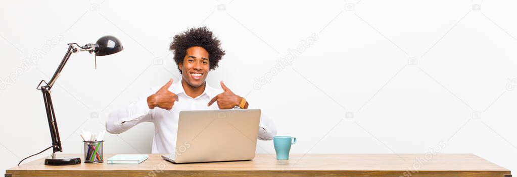 young black businessman feeling happy, surprised and proud, pointing to self with an excited, amazed look on a desk