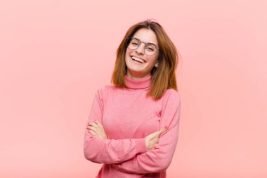 Genç ve güzel bir kadın kollarını kavuşturup mutlu bir şekilde gülüyor. Pembe arka planda rahat, pozitif ve tatmin edici bir poz veriyor.