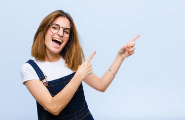 Genç, güzel bir kadın sevinçli ve şaşırmış hissediyor. Şok olmuş bir ifadeyle gülümsüyor ve mavi arka plana işaret ediyor.