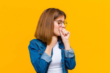 Gırtlağı ağrıyan ve grip belirtileri gösteren, ağzı sarı arka planda öksüren genç ve güzel bir kadın.