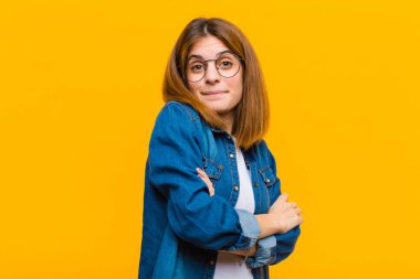 Genç, güzel bir kadın omuz silkiyor, kafası karışık ve kararsız, kolları bağlı ve şaşkın, sarı arka planda.