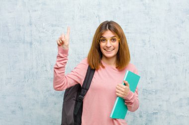 Genç bayan öğrenci neşeyle ve mutlu bir şekilde gülümsüyor, bir eliyle yukarı işaret ederek grunge duvarının arka planına alanı kopyalıyor.